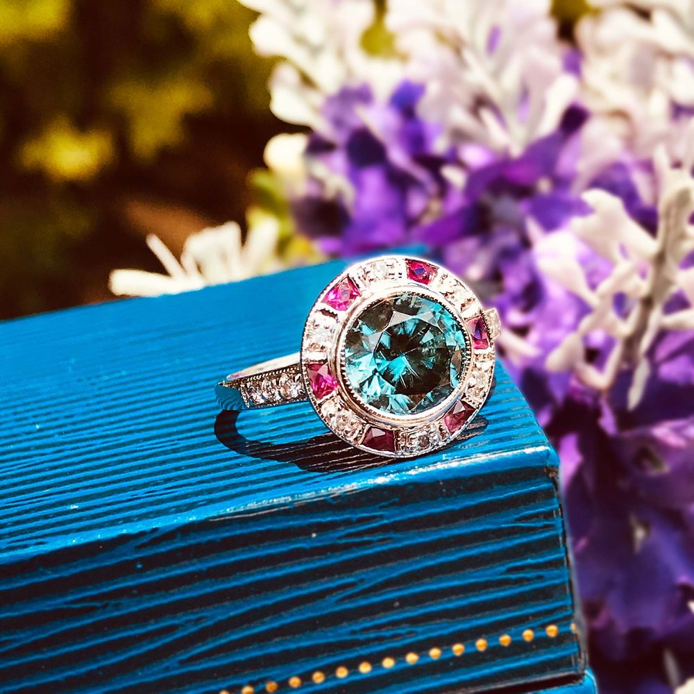 2.75ct Blue Zircon Ring with Ruby and Diamond Halo Cluster, central round-cut blue zircon is surrounded by rubies with a deep pink hue and diamonds with diamond-set shoulders in 14ct white gold