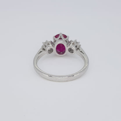 An oval ruby and brilliant cut diamond three stone ring in white metal, against a white background