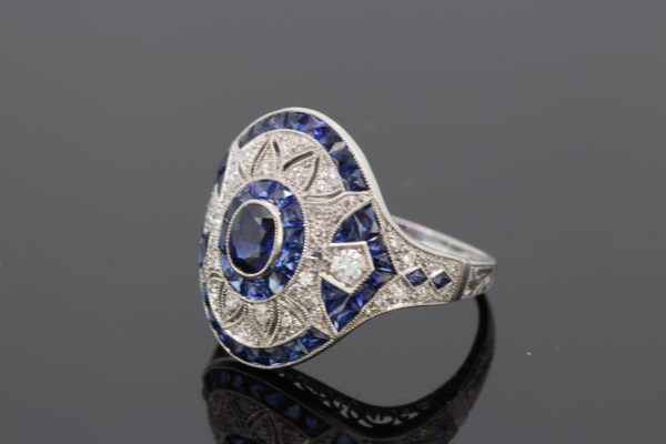 Art Deco Style Sapphire and Diamond Platinum Dress Ring; central oval faceted sapphire within diamond set pierced surround and calibre-cut sapphire outer border