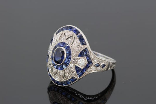 Art Deco Style Sapphire and Diamond Platinum Dress Ring; central oval faceted sapphire within diamond set pierced surround and calibre-cut sapphire outer border