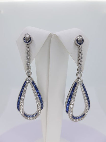 A pair of sapphire and diamond teardrop shaped long drop earrings in white gold, on an earring display pad, against a white background