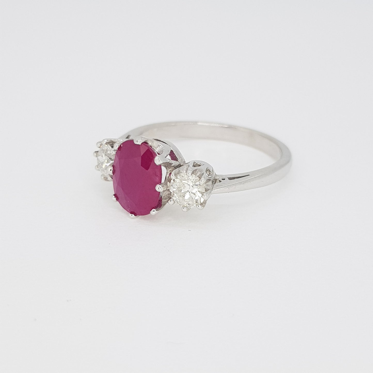 An oval ruby and brilliant cut diamond three stone ring in white metal, against a white background