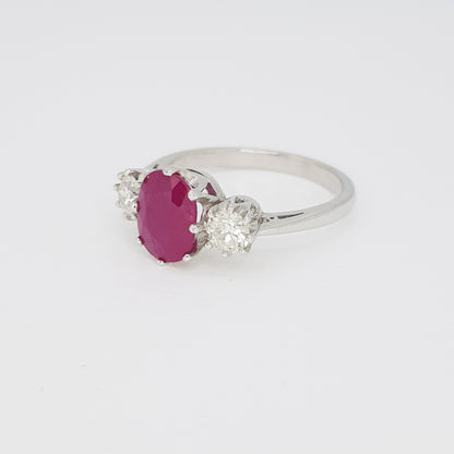An oval ruby and brilliant cut diamond three stone ring in white metal, against a white background