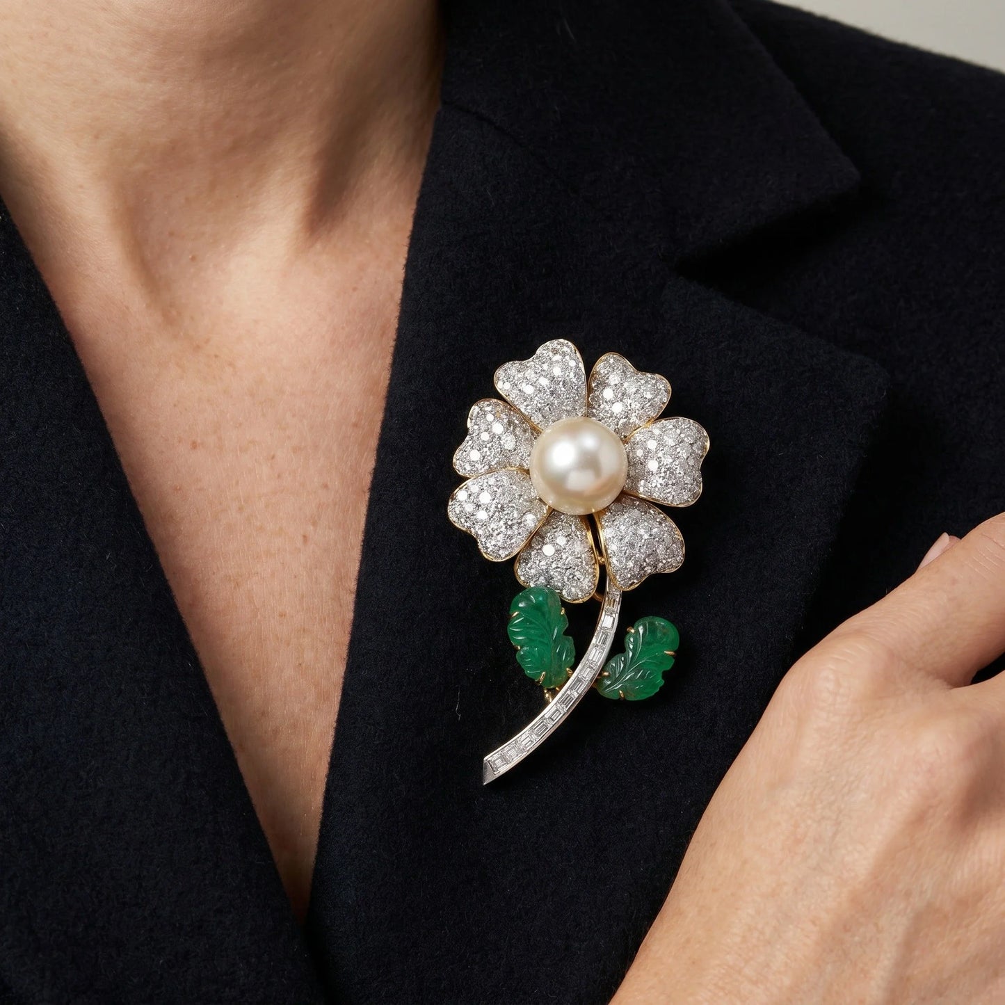 A flower brooch set with diamonds, a pearl centre and emerald leaves, in yellow and white gold, modelled on a black jacket