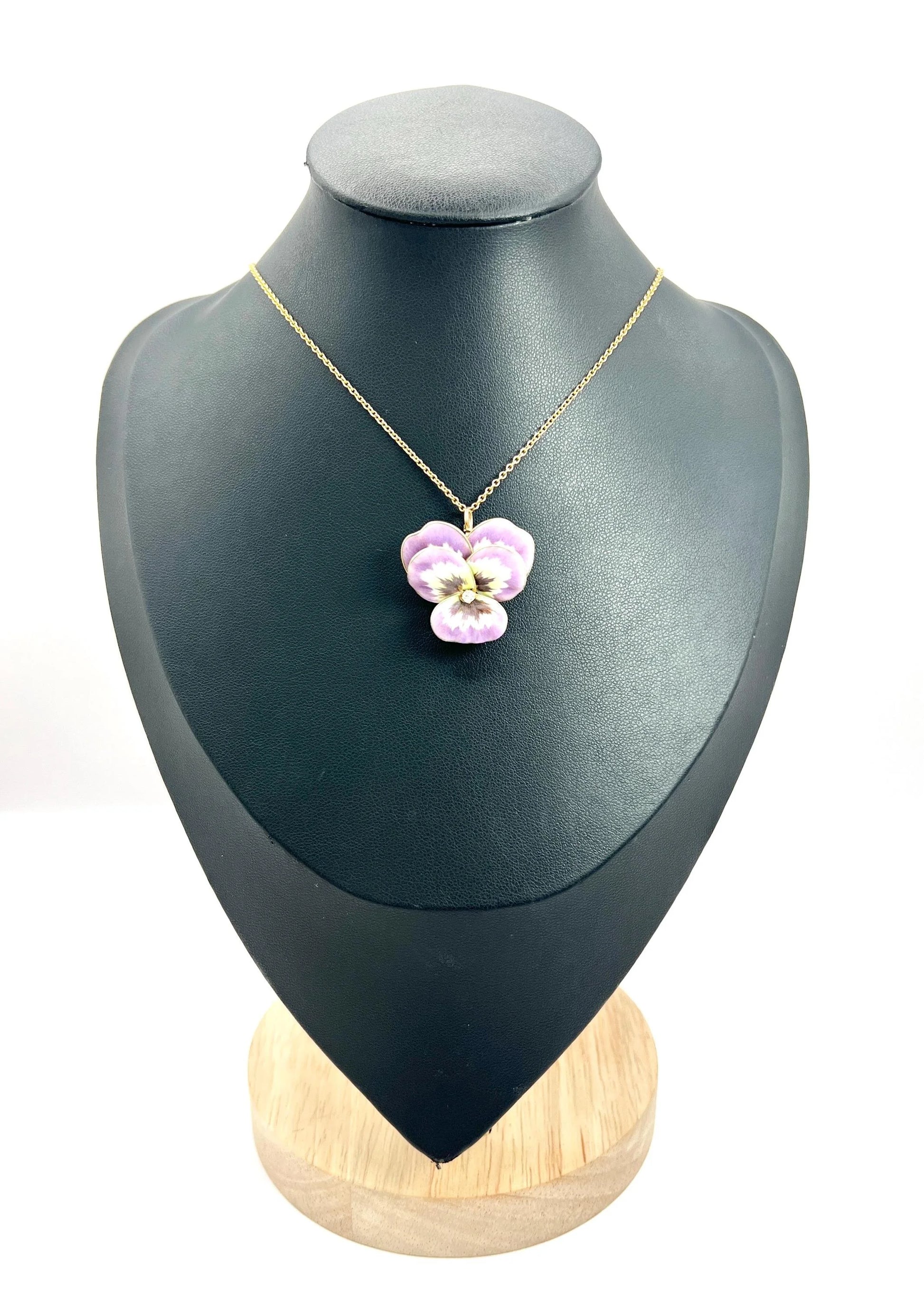 An enamelled pansy flower pendant/brooch with a diamond centre, in yellow gold, with a chain, on a black jewellery bust