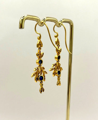 A pair of sapphire and diamond foliate drop earrings in yellow gold, hung on a hook