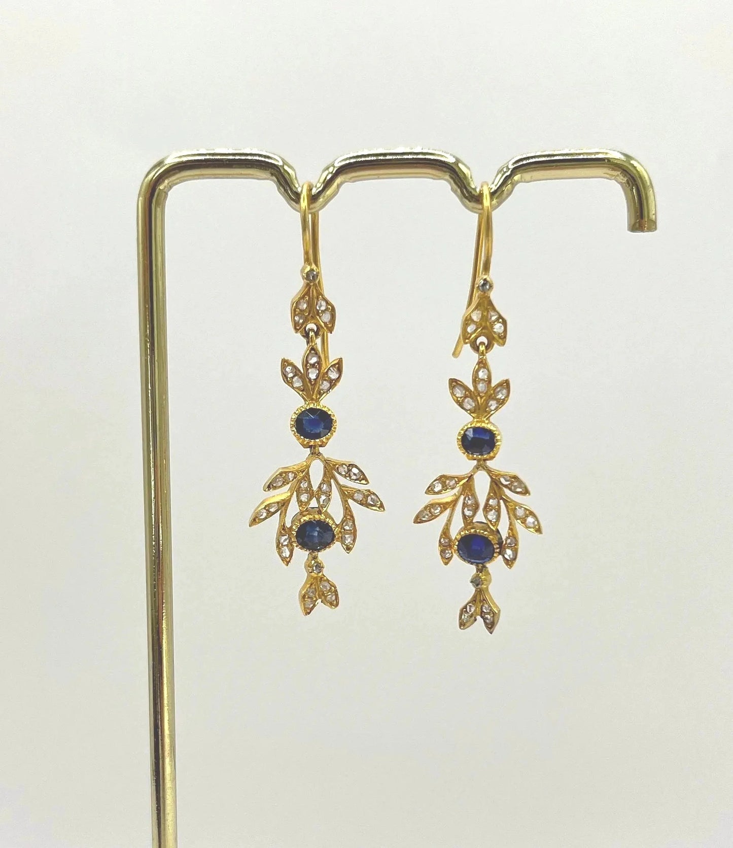 A pair of sapphire and diamond foliate drop earrings in yellow gold, hung on a hook