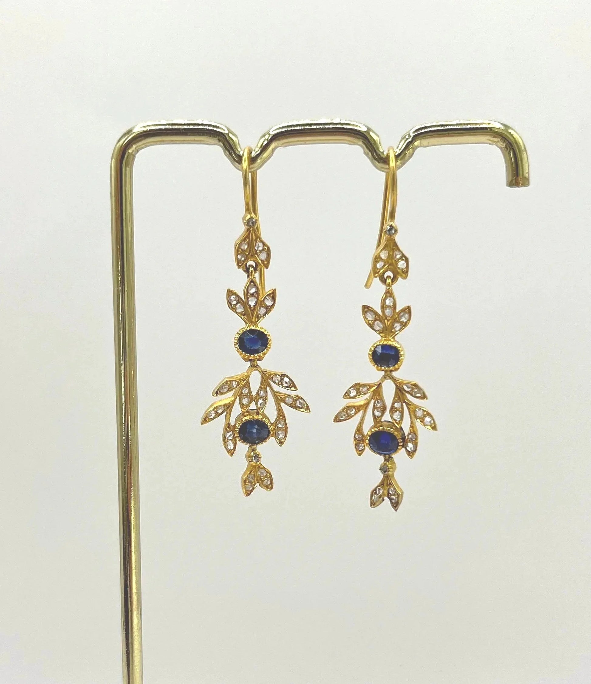 A pair of sapphire and diamond foliate drop earrings in yellow gold, hung on a hook