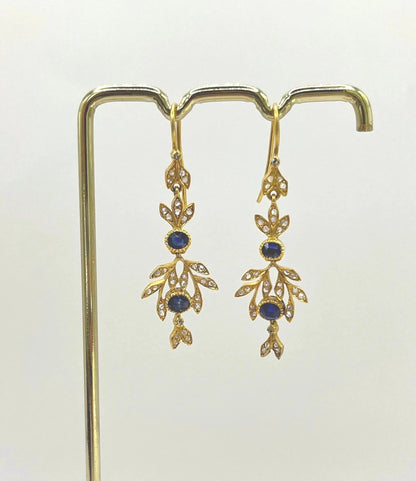 A pair of sapphire and diamond foliate drop earrings in yellow gold, hung on a hook