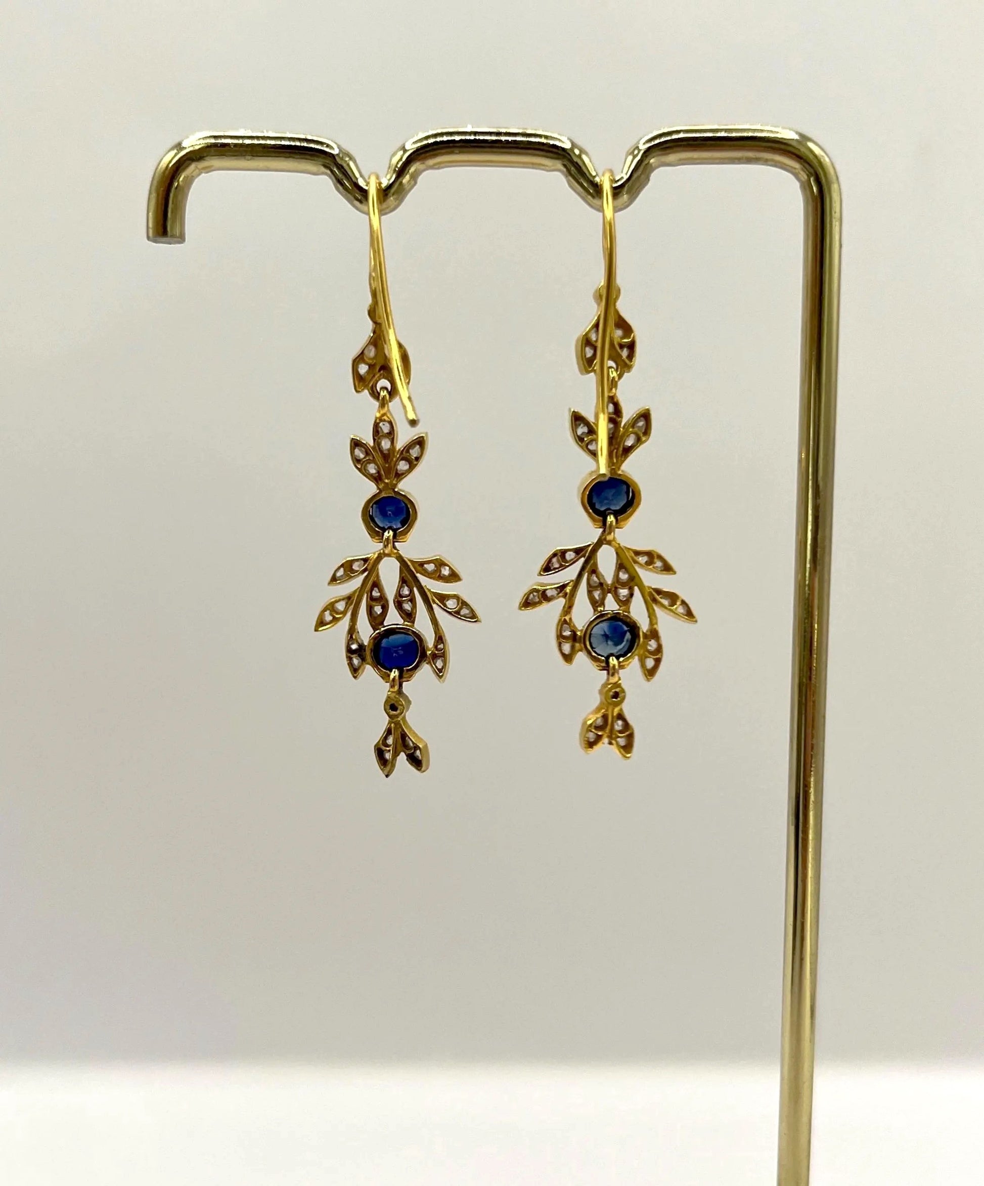 A pair of sapphire and diamond foliate drop earrings in yellow gold, hung on a hook