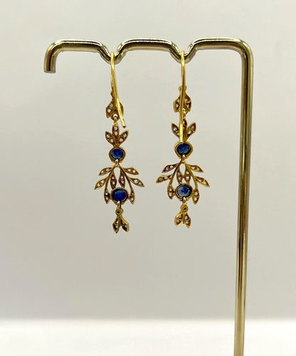 A pair of sapphire and diamond foliate drop earrings in yellow gold, hung on a hook