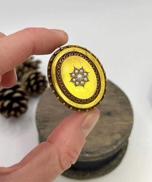 An oval yellow gold brooch with a seed pearl cluster, held in a hand