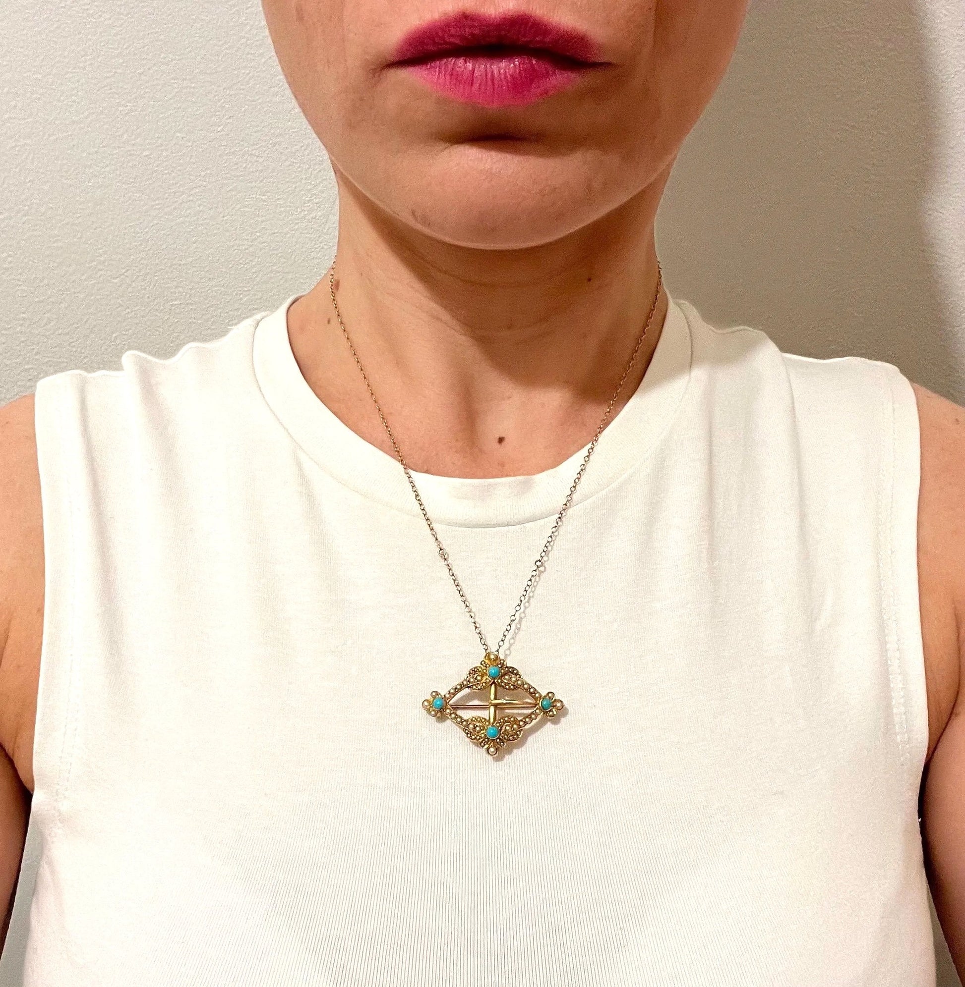 A yellow gold buckle style brooch/pendant with turquoise and seed pearls, with a chain, modelled by a woman wearing a white top