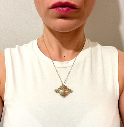 A yellow gold buckle style brooch/pendant with turquoise and seed pearls, with a chain, modelled by a woman wearing a white top
