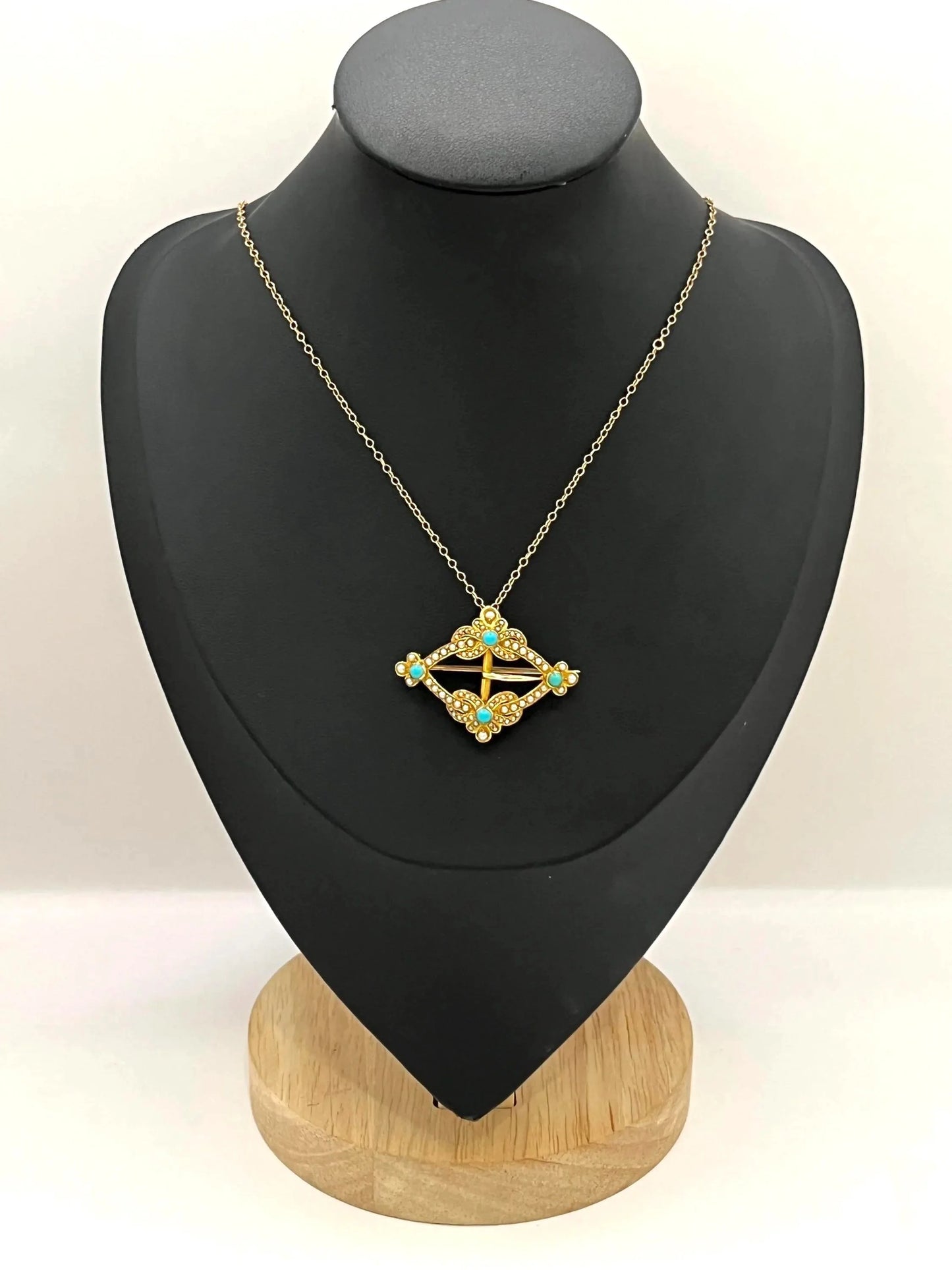 A yellow gold buckle style brooch/pendant with turquoise and seed pearls, with a chain, on a black jewellery bust