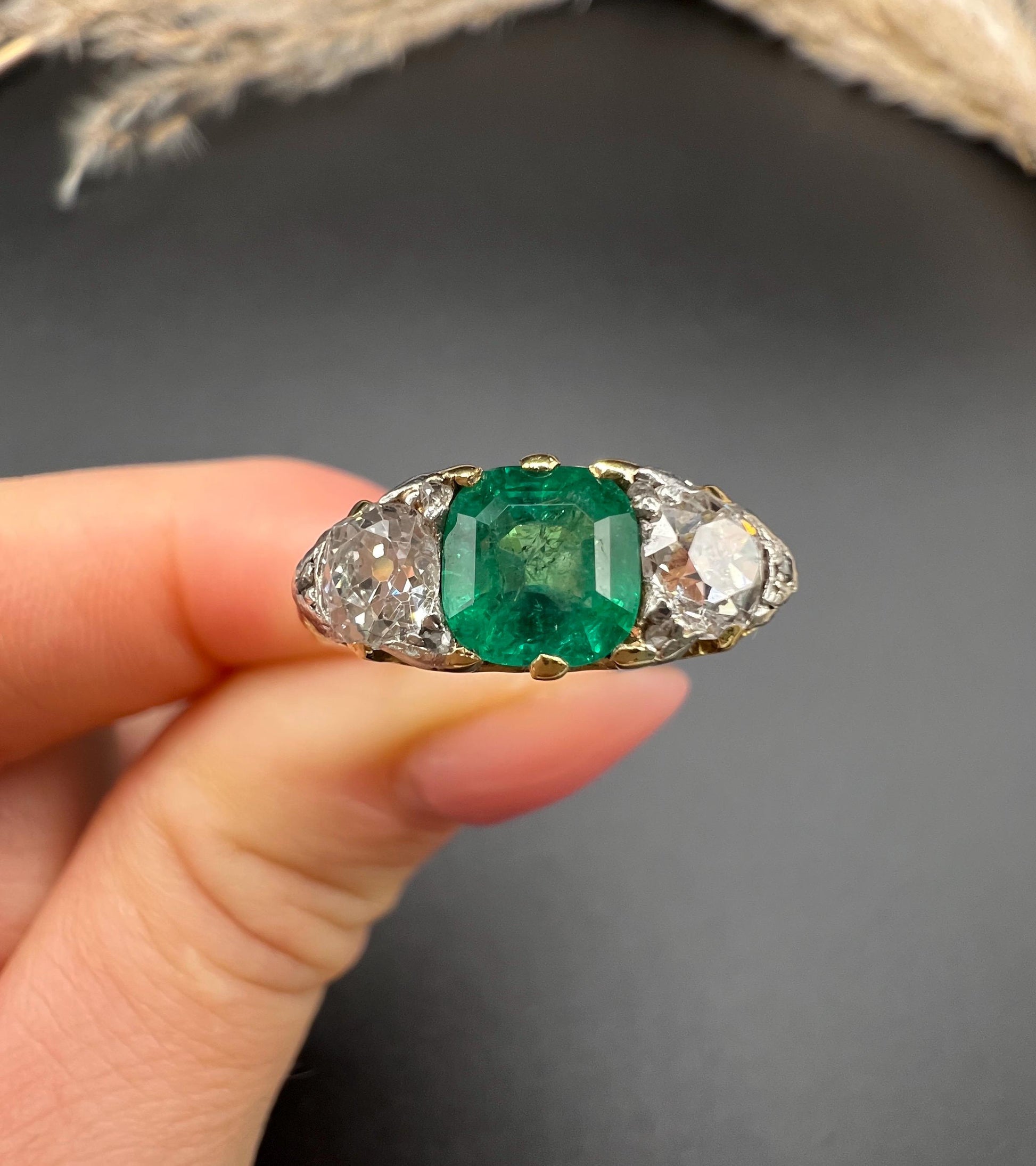 A three stone old cut emerald and diamond ring in yellow gold, held in a hand