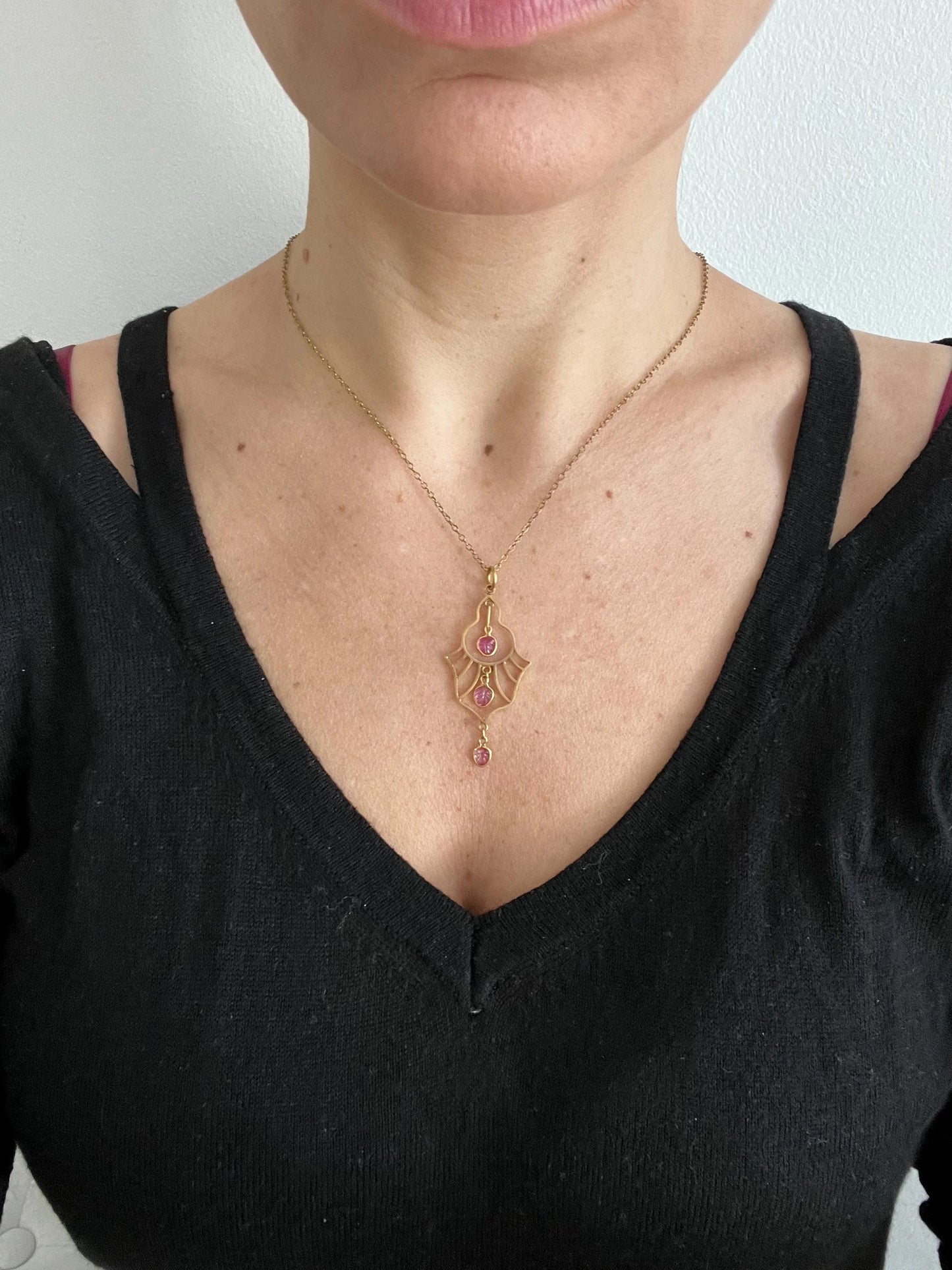 A yellow gold three stone ruby openwork drop pendant on a yellow gold chain modelled by a woman wearing a black top