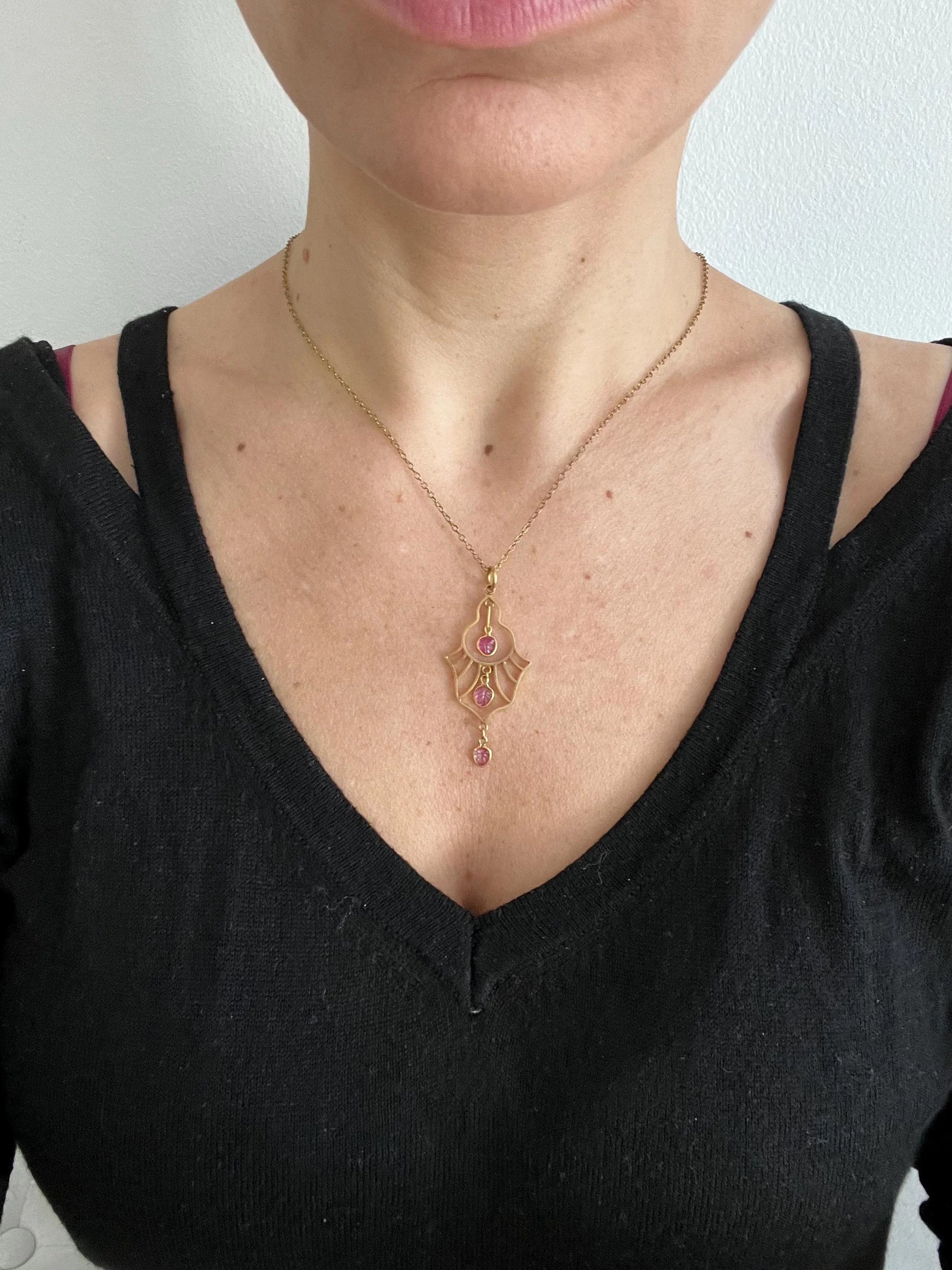 A yellow gold three stone ruby openwork drop pendant on a yellow gold chain modelled by a woman wearing a black top