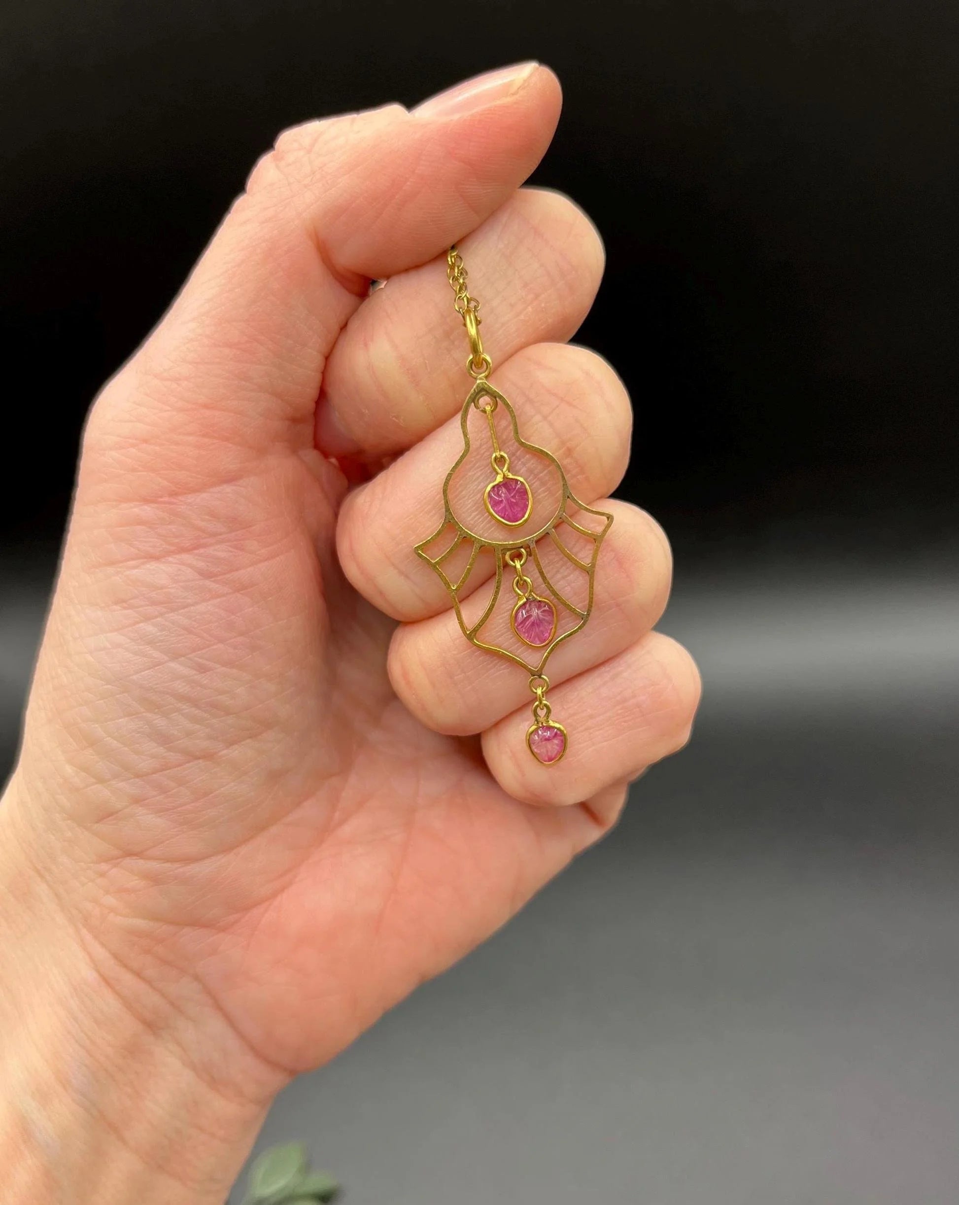 A yellow gold three stone ruby openwork drop pendant on a yellow gold chain, held in a hand
