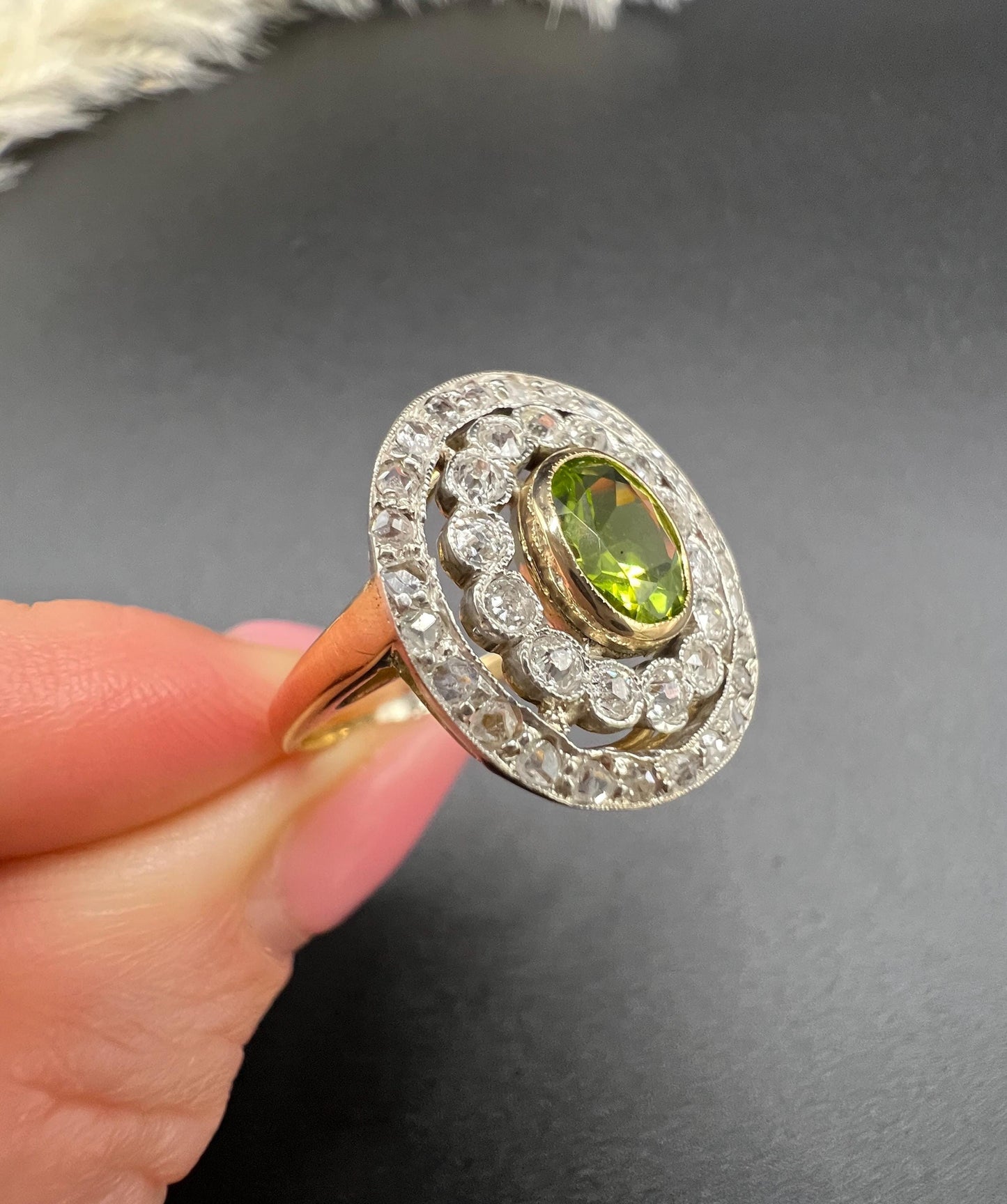An oval peridot and old cut diamond fancy cluster ring in yellow gold and platinum held in a hand
