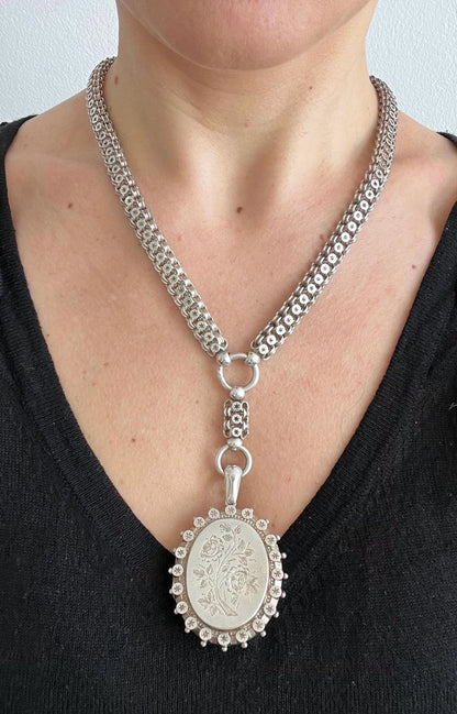A silver oval locket with floral engraving on a silver book chain collar, modelled