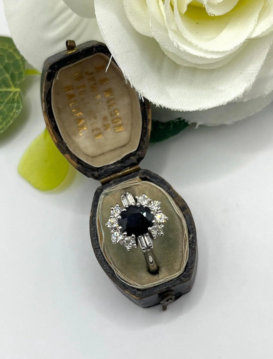 An oval cluster ring with a sapphire centre, round diamond surround and baguette cut diamond shoulders in white gold, in an antique ring box