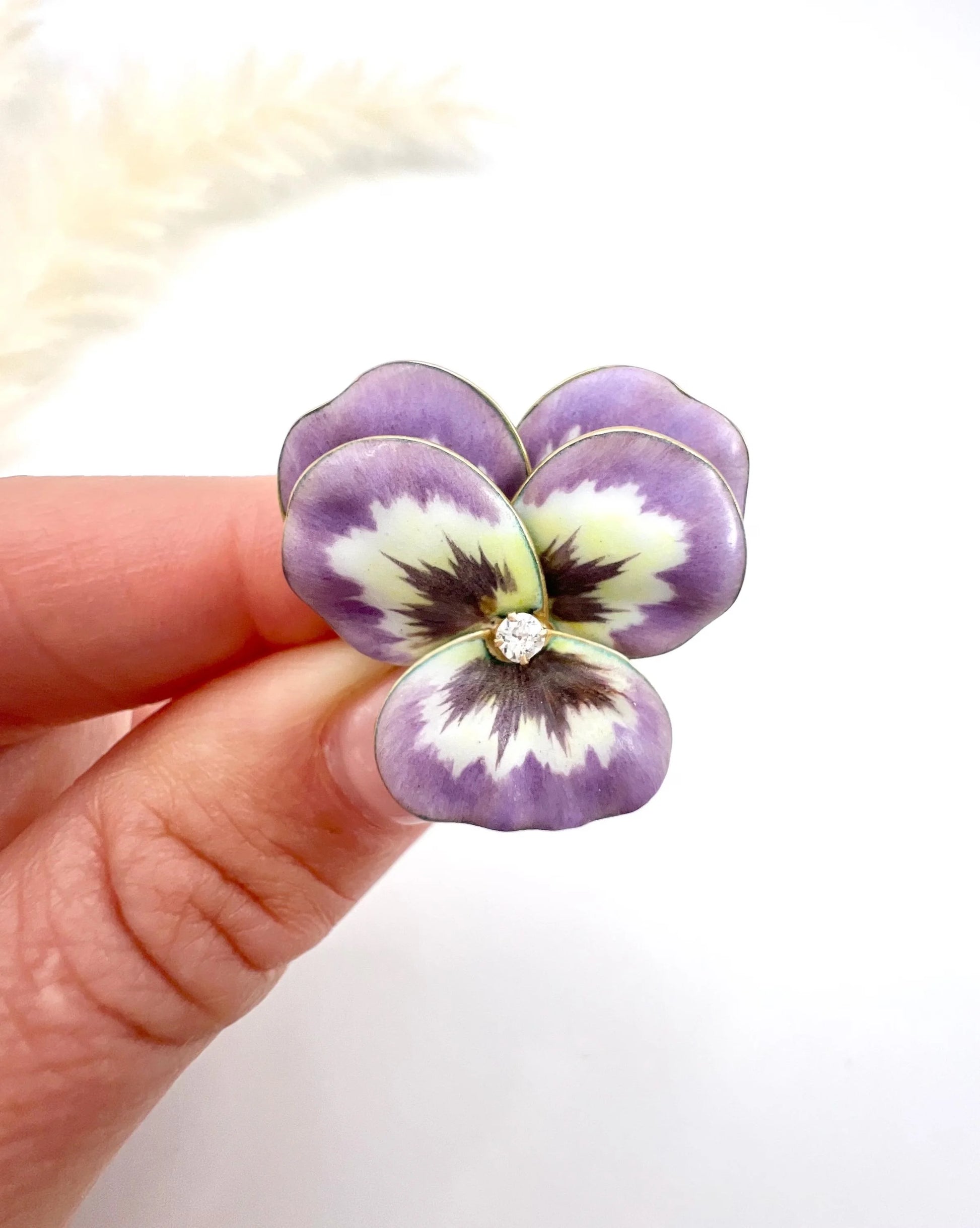 An enamelled pansy flower pendant/brooch with a diamond centre, in yellow gold, held in a hand