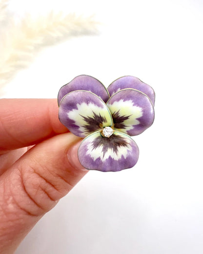 An enamelled pansy flower pendant/brooch with a diamond centre, in yellow gold, held in a hand