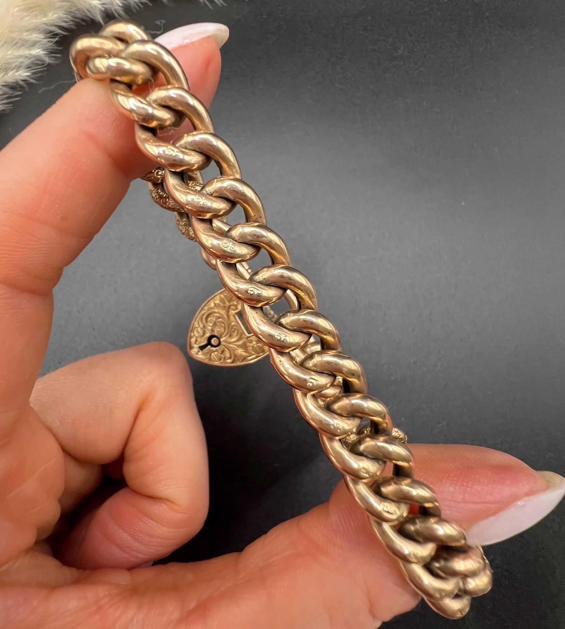 A rose gold curb link bracelet with engraved heart padlock clasp, held in a hand