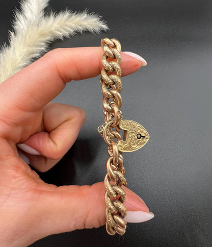 A rose gold curb link bracelet with engraved heart padlock clasp, held in a hand