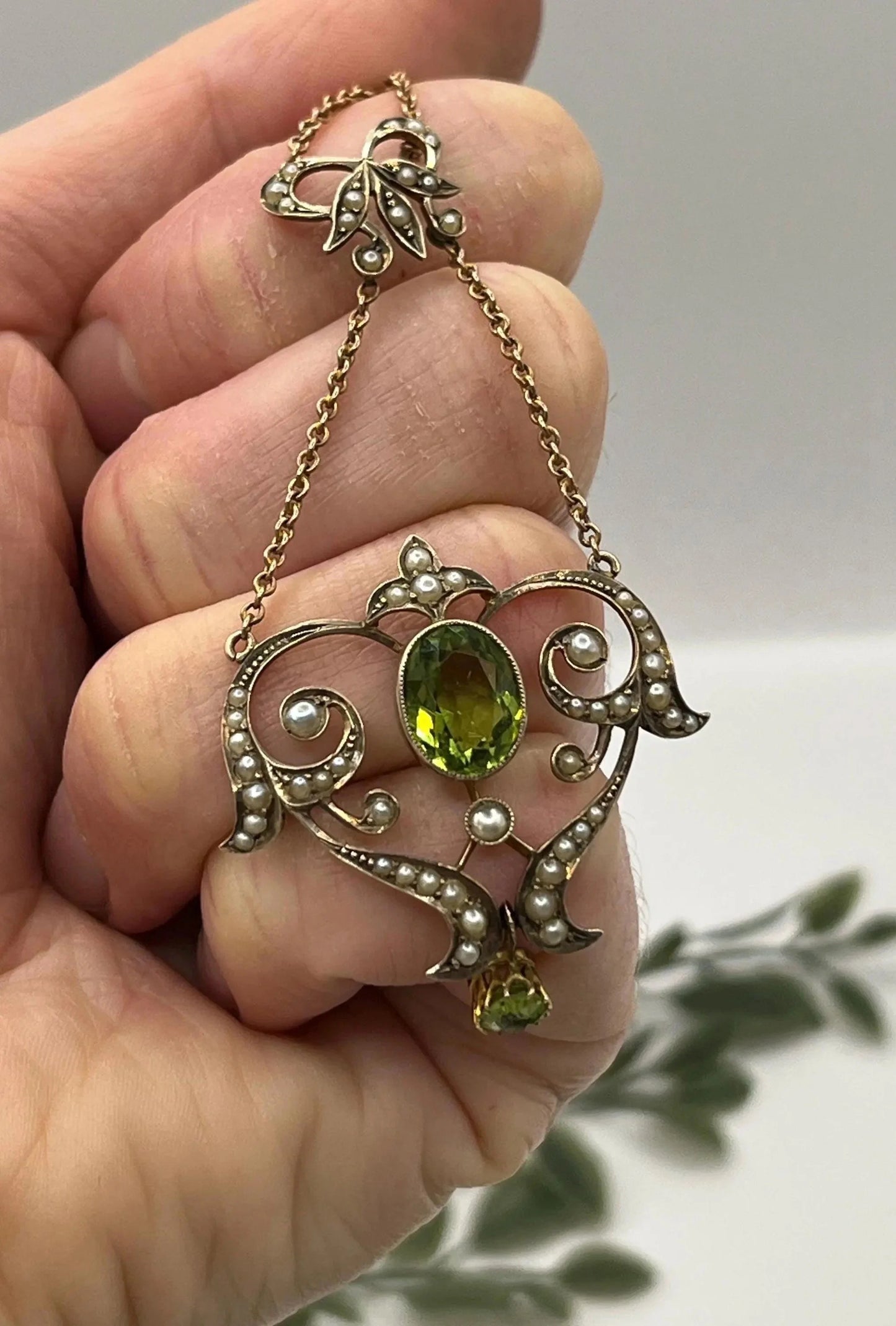 A yellow gold peridot and seed pearl pendant necklace, held in a hand