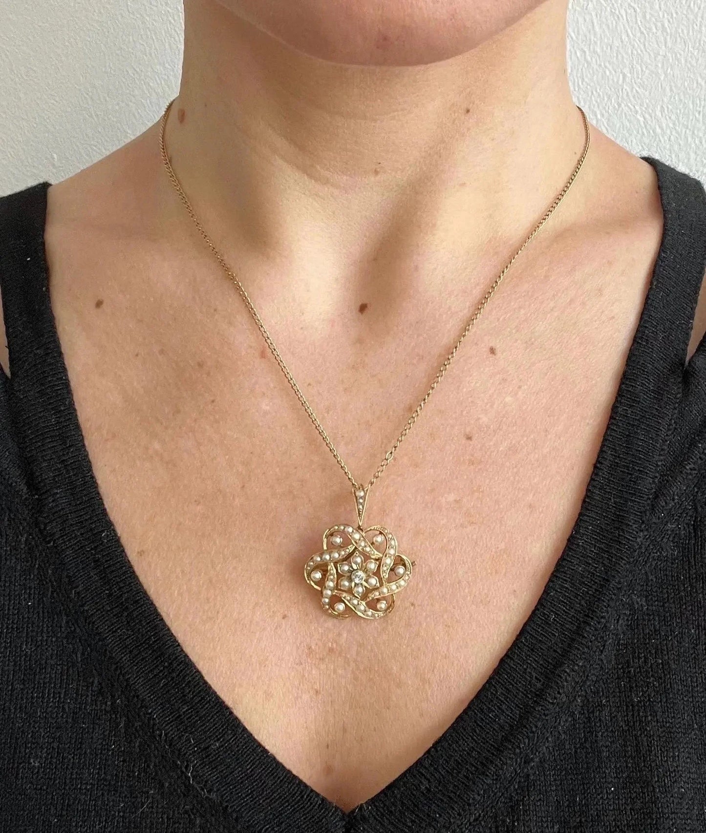 A yellow gold seed pearl and diamond floral brooch/pendant, with a chain, modelled by a woman wearing a black top