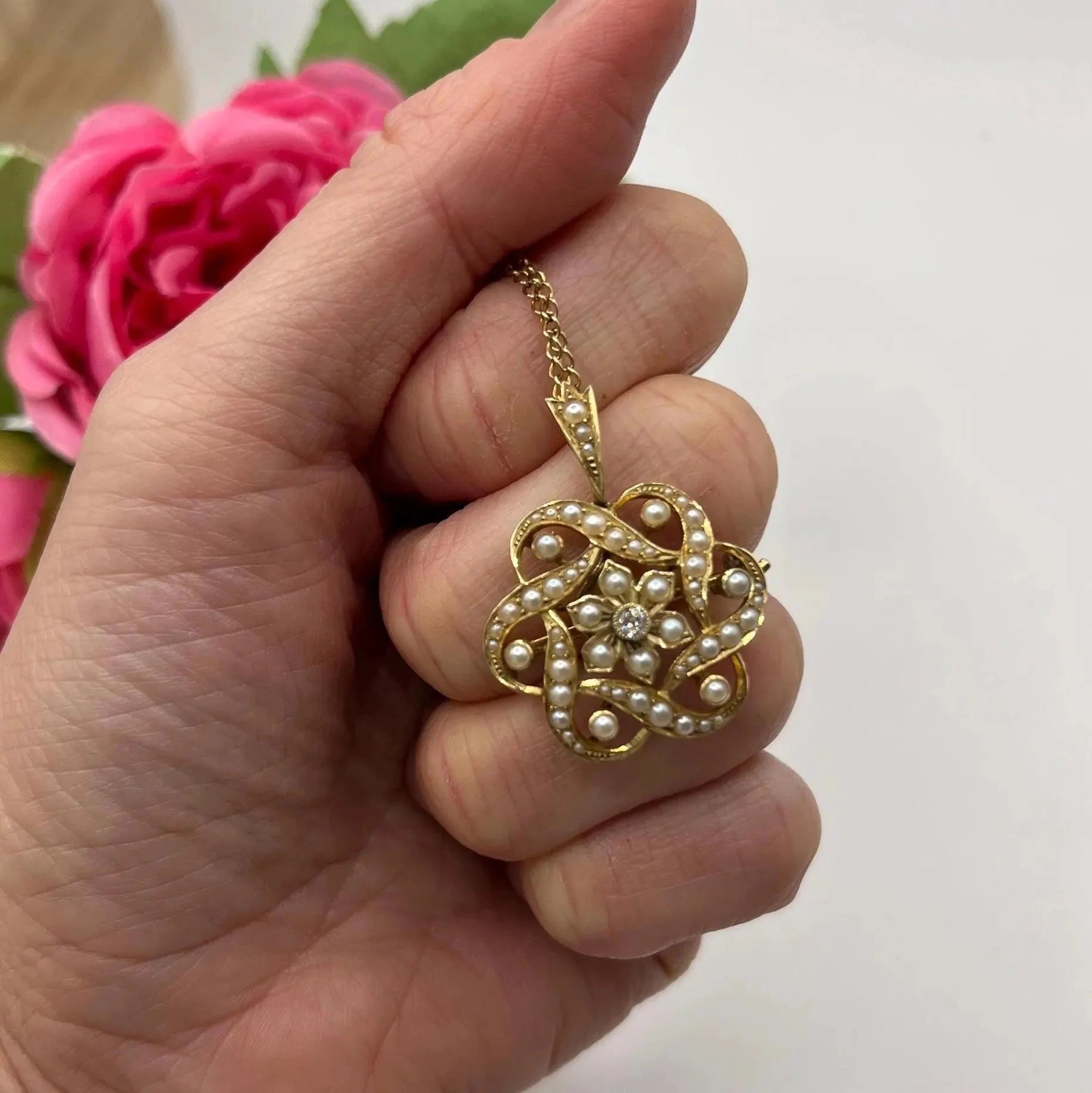 A yellow gold seed pearl and diamond floral brooch/pendant, with a chain, held in a hand