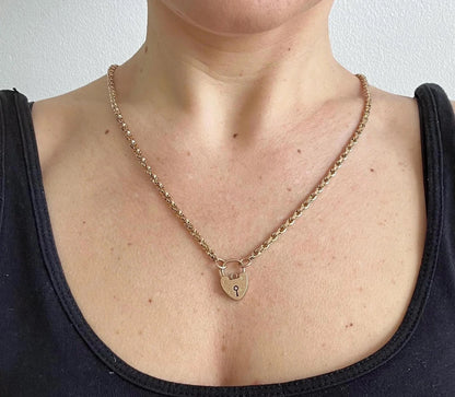 A yellow gold chain necklace with a heart shaped padlock clasp, modelled by a woman wearing a black top