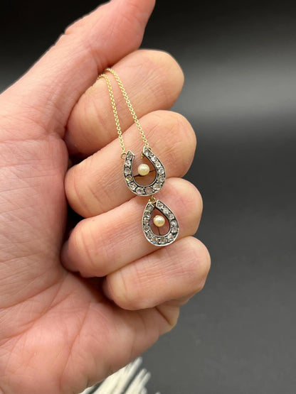 A pearl and diamond set doube horseshoe pendant on a gold chain, held in a hand