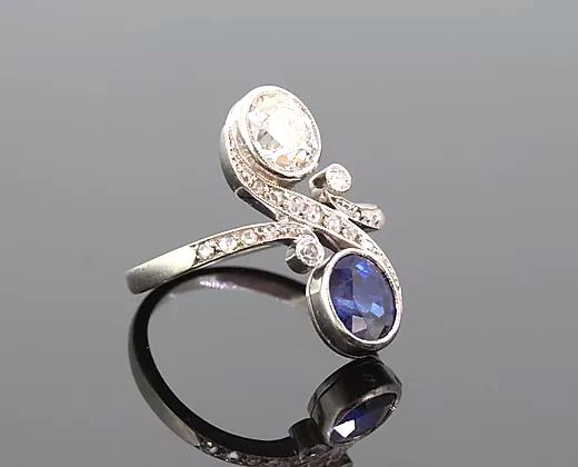 Art Nouveau Sapphire and Diamond Crossover Ring; oval-cut sapphire and oval-cut diamond, accented with diamond set swirls and shoulders, 18ct white gold.