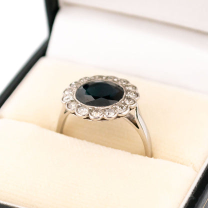 A flower shaped cluster ring set with a central sapphire surrounded by a halo of round diamonds, in platinum, in a ring box