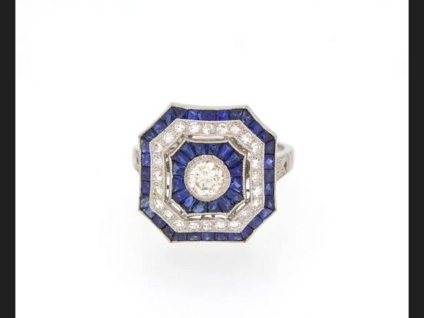 Art Deco Style Sapphire and Diamond Calibre Set Ring; Central round cut diamond encompassed by baguette cut sapphires and round cut diamonds, set in platinum