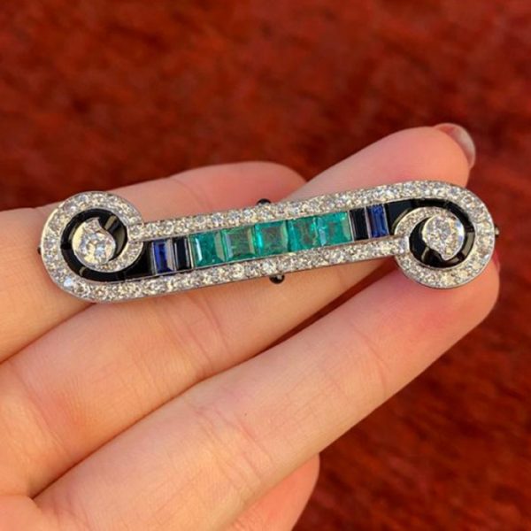 Art Deco French Emerald Sapphire Onyx and Diamond Bar Brooch in Platinum; set with 2.00cts calibre-cut emeralds, 0.50cts calibre-cut sapphires and fancy-cut black onyx, framed by 2.50cts old-cut and single-cut diamonds, in a scroll design, with enamelled detail
