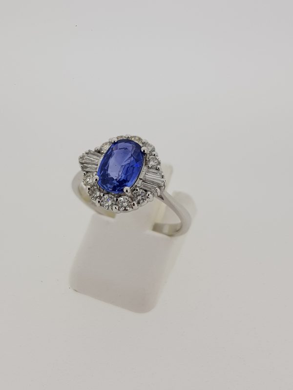 Modern 1.67ct Sapphire and Diamond Cluster Ring in 18ct White Gold; 1.67 carat oval faceted sapphire surrounded by brilliant-cut diamonds with tapered baguette-cut diamond set to the shoulders
