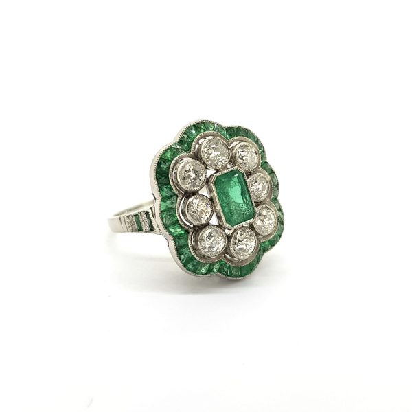 Art Deco Style Emerald and Diamond Cluster Dress Ring; central emerald-cut emerald surrounded by brilliant cut diamonds, within a border of calibre cut emeralds, in platinum