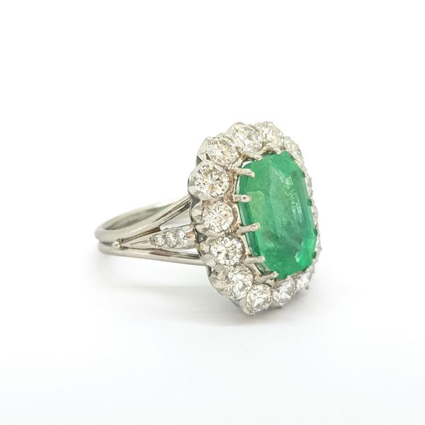 Vintage Emerald and Diamond Cluster Ring; 5ct emerald-cut emerald within a cluster of 2.50cts round brilliant-cut diamonds, in 18ct white gold, with openwork under-gallery, split shoulders with a central diamond set post