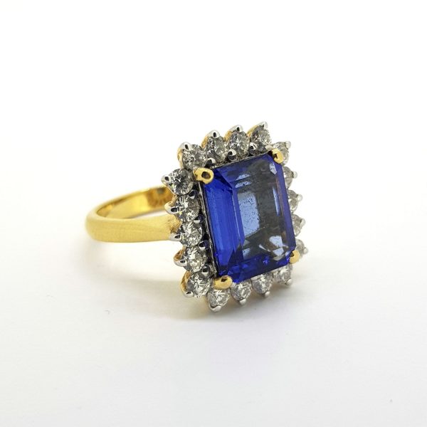 Emerald Cut Tanzanite and Diamond Cluster Ring; central 4.45ct emerald-cut tanzanite within a 1.04ct diamond surround, in 18ct yellow gold