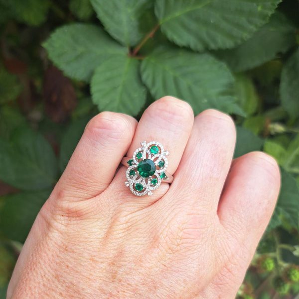 Decorative 1.13ct Emerald and Diamond Cluster Dress Ring, 1.13 carat emerald nestled within entwinned loop diamond surround with emerald accents in 18ct white gold