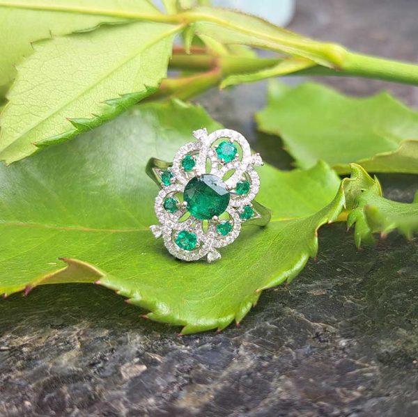 Decorative 1.13ct Emerald and Diamond Cluster Dress Ring, 1.13 carat emerald nestled within entwinned loop diamond surround with emerald accents in 18ct white gold