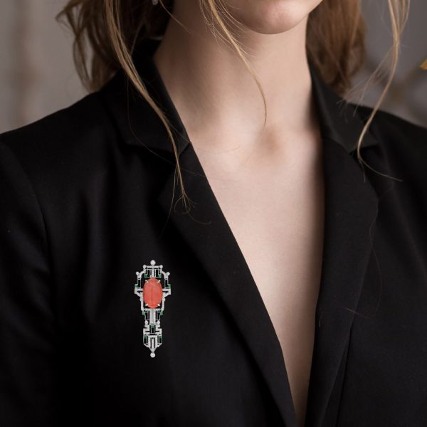 Art Deco Inspired Coral Onyx and Diamond Brooch, vivid orange 6.32ct oval coral within diamond geometric frame with contrasting black onyx and green emerald accents in 14ct white gold