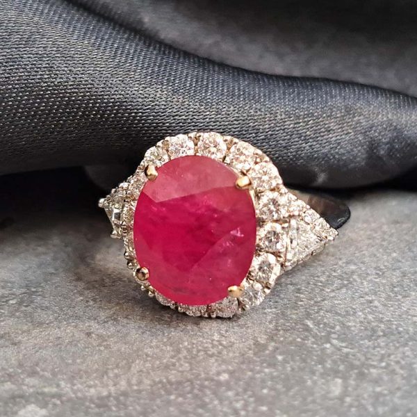 Contemporary 4.79ct Ruby and Diamond Cluster Engagement Ring, 4.79ct oval red ruby flanked by trillion diamonds all within a brilliant-cut diamond halo border in 18ct white gold