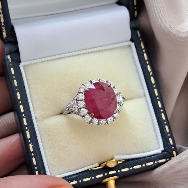 Contemporary 4.79ct Ruby and Diamond Cluster Engagement Ring, 4.79ct oval red ruby flanked by trillion diamonds all within a brilliant-cut diamond halo border in 18ct white gold