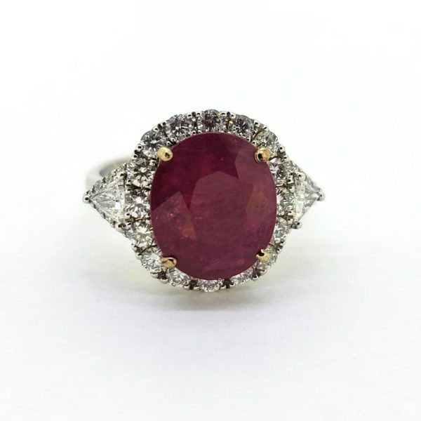 Contemporary 4.79ct Ruby and Diamond Cluster Engagement Ring, 4.79ct oval red ruby flanked by trillion diamonds all within a brilliant-cut diamond halo border in 18ct white gold