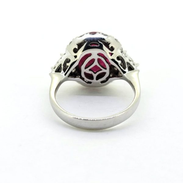Contemporary 4.79ct Ruby and Diamond Cluster Engagement Ring, 4.79ct oval red ruby flanked by trillion diamonds all within a brilliant-cut diamond halo border in 18ct white gold